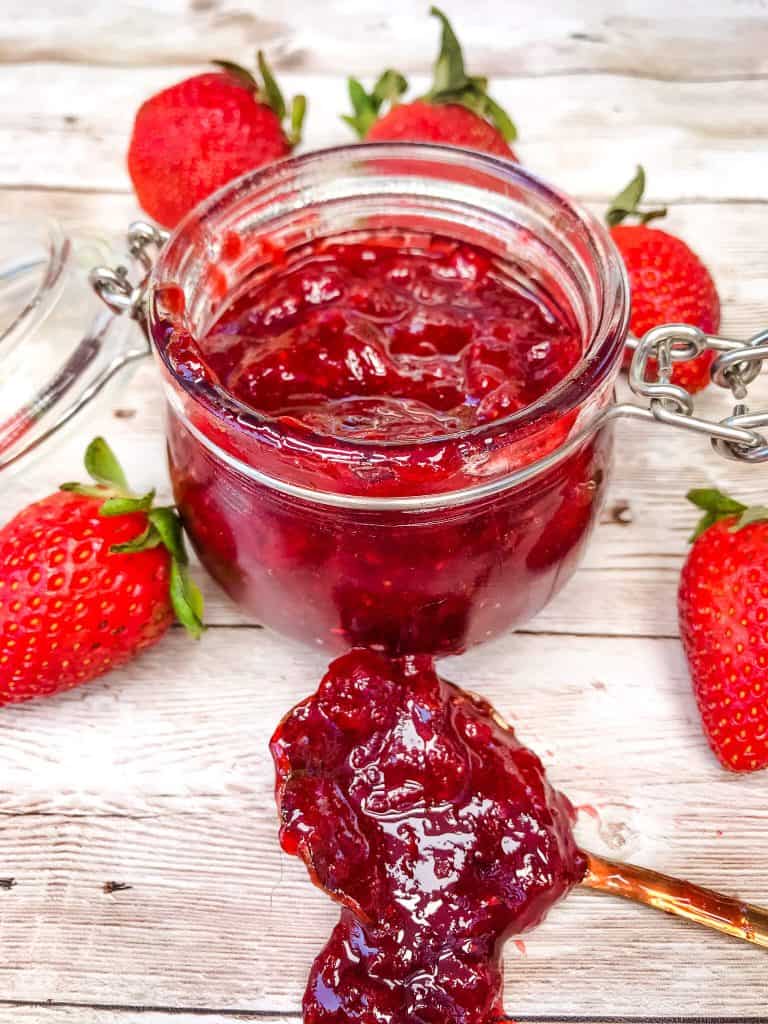 Small clear jar of Strawberry Jam surrounded by strawberries with a copper spoon in front that has jam on it