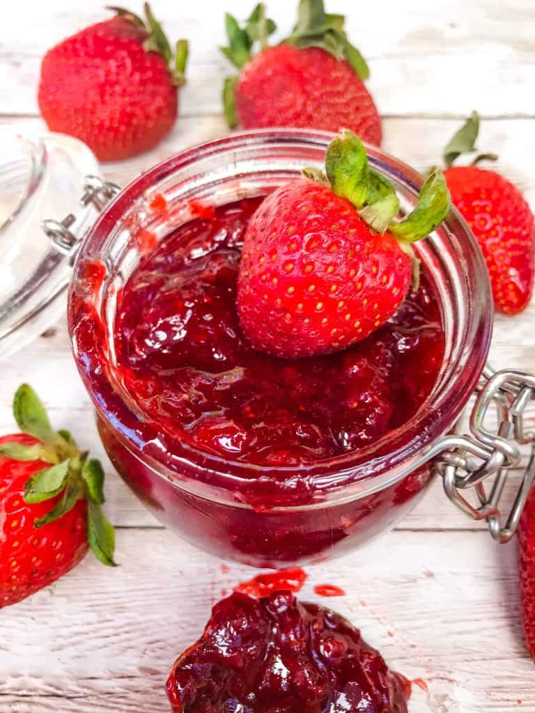 Top view of a small clear jar filled with homemade strawberry jam and a strawberry on top, surrounded by strawberries on wood planks