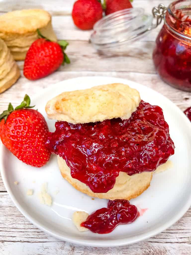 How to Make Homemade Strawberry Jam on a fresh biscuit with the top on, on a small white plate with a strawberry next to it