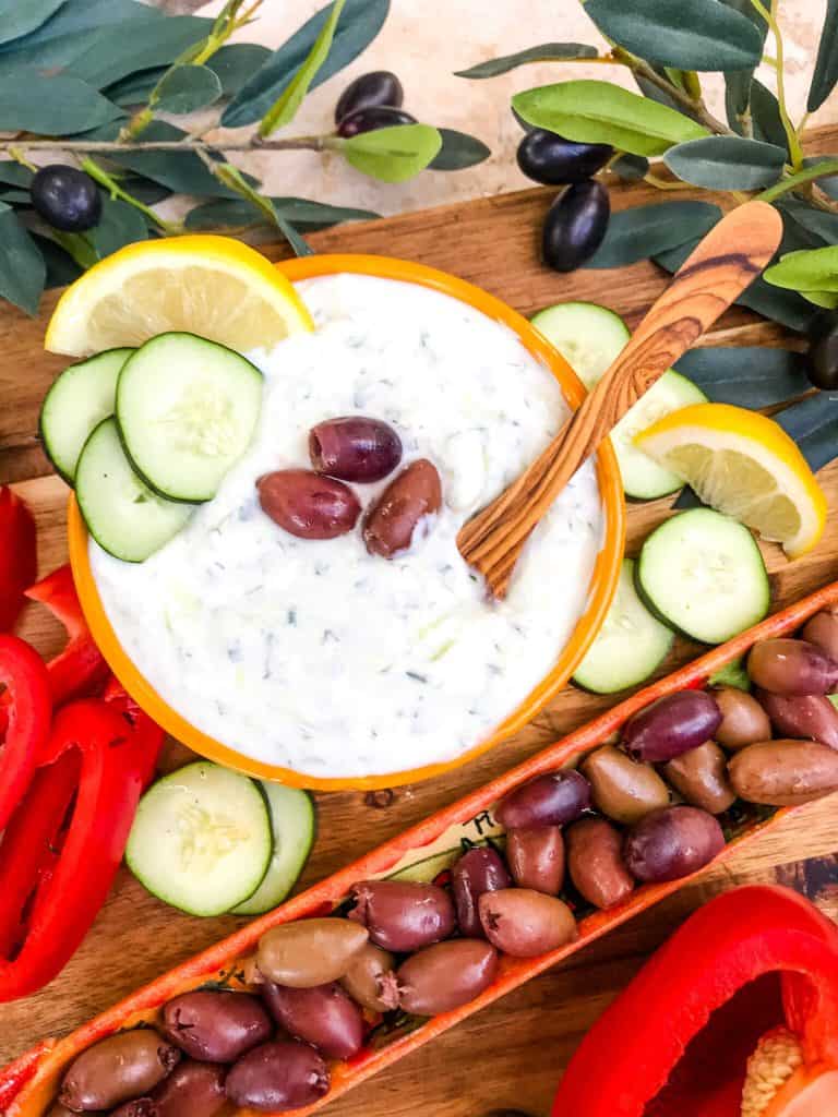 A bowl of Greek Tzatziki Yogurt Sauce with a wooden spoon surrounded by cucumber, lemon, and olives.