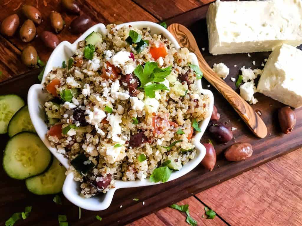 A bowl of Greek Quinoa Salad surrounded by ingredients on wood boards