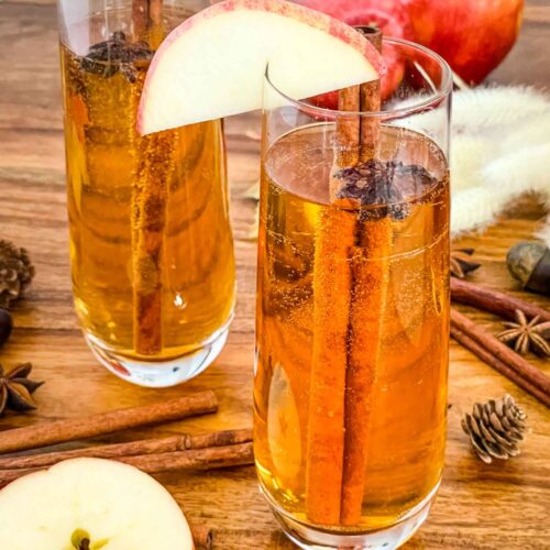 Two flutes of Cinnamon Apple Cider Mimosa on a wood table with apple slices and dried spices.
