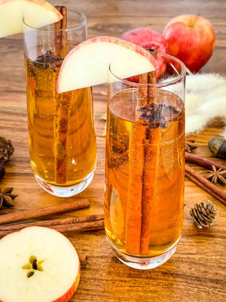 Two flutes of Cinnamon Apple Cider Mimosa on a wood table with apple slices and dried spices.