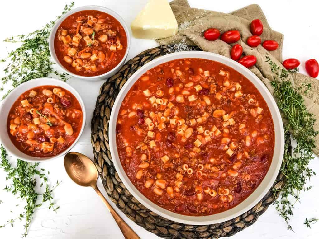 A large bowl and two serving sized bowls of Copycat Olive Garden Pasta Fagioli Soup surrounded by herbs