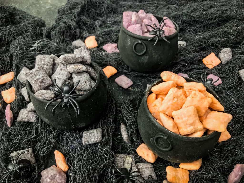 Cauldrons filled with separated orange, black, and purple Halloween Puppy Chow on a gray gauze