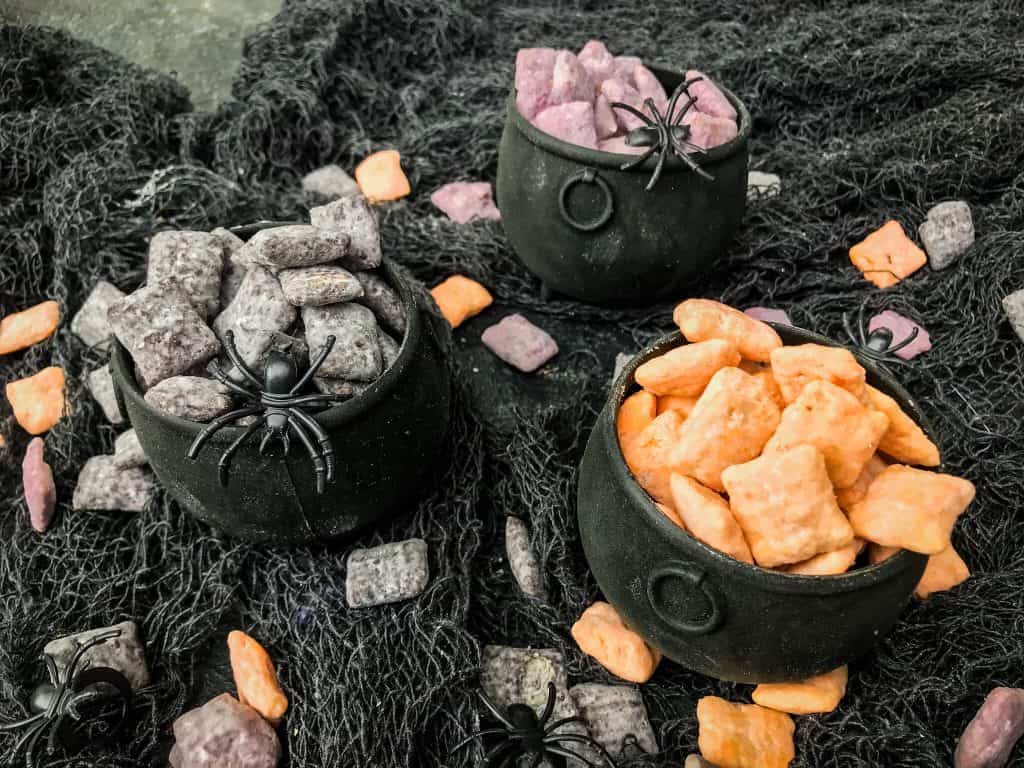 Cauldrons filled with separated orange, black, and purple Halloween Puppy Chow on a gray gauze