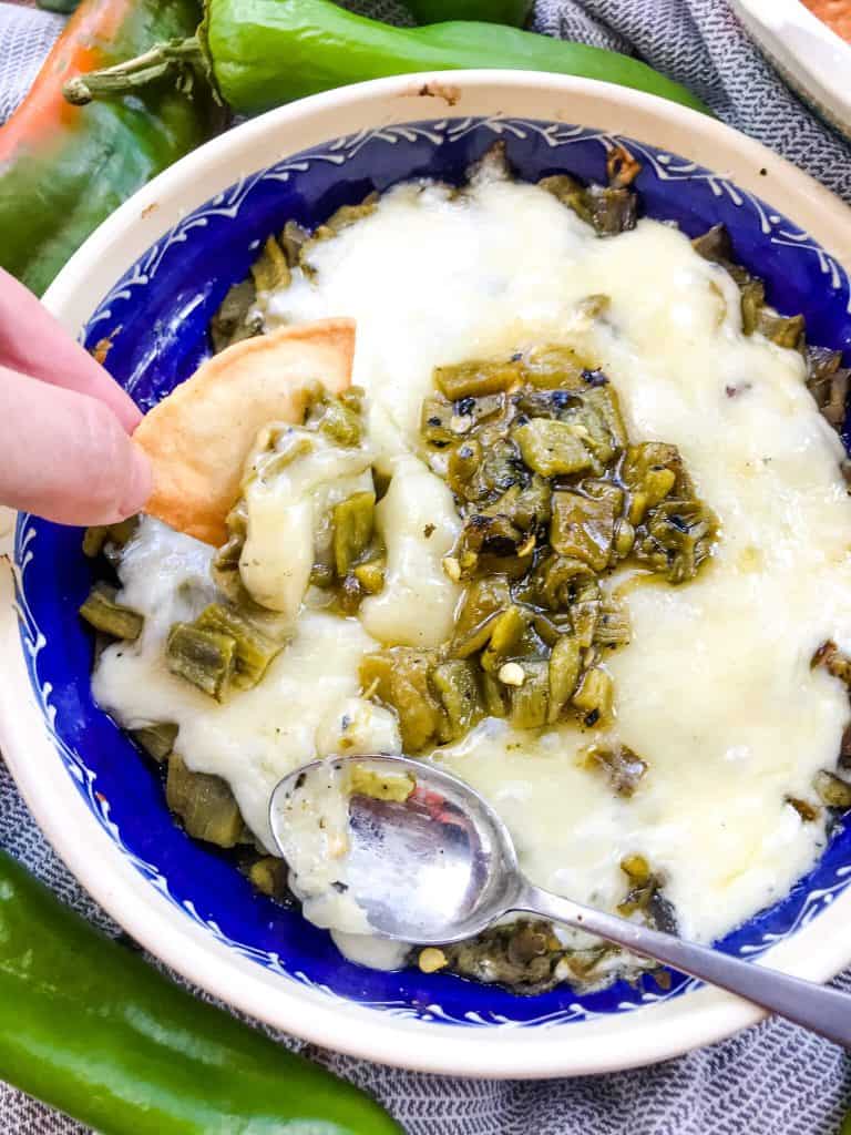 A hand dipping a chip into melted cheese with Hatch peppers