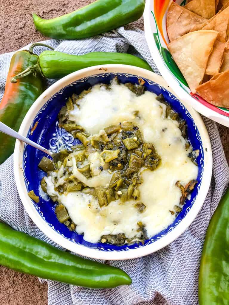 A spoon in a dish of Hatch Green Chile Queso Fundido on a towel surrounded by peppers and chips