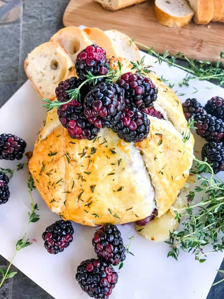 A full Thyme Blackberry Baked Brie in Puff Pastry on a marble board surrounded by thyme and berries
