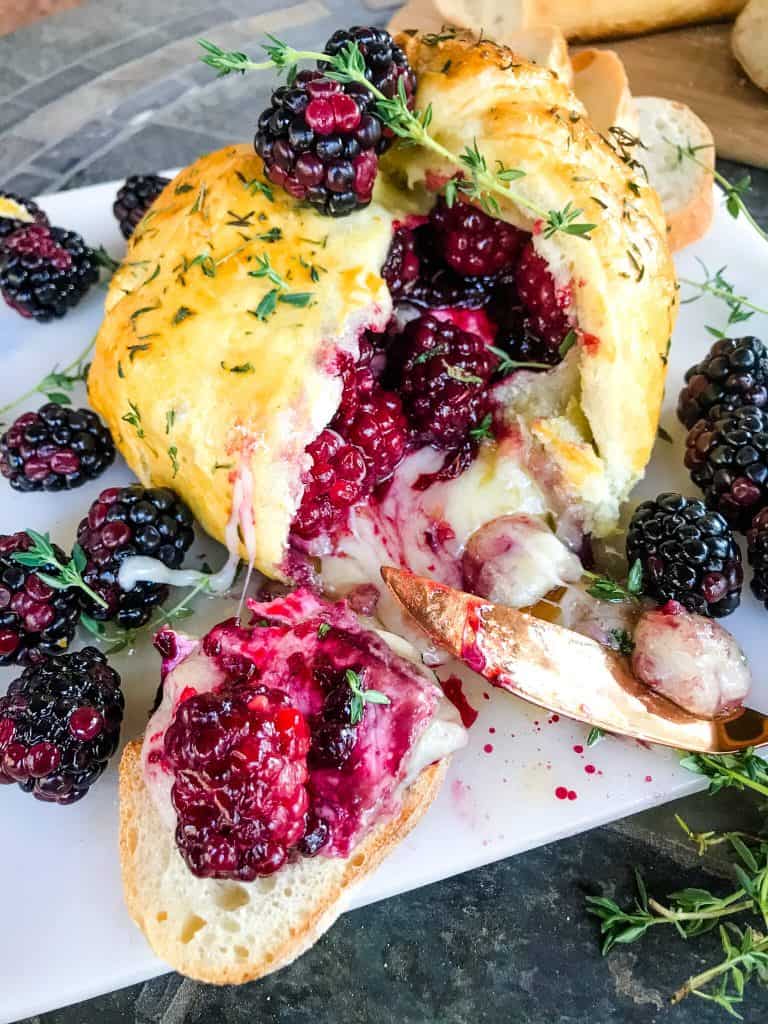 Thyme Blackberry Baked Brie in Puff Pastry cut open with cheese oozing out surrounded by berries and crostini