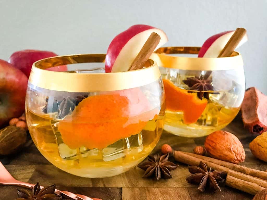 Two round cocktail glasses filled with an orange peel, apple, cinnamon, and the drink