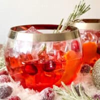 A glass of Cranberry Old Fashioned Cocktail with cranberries and rosemary in and around it