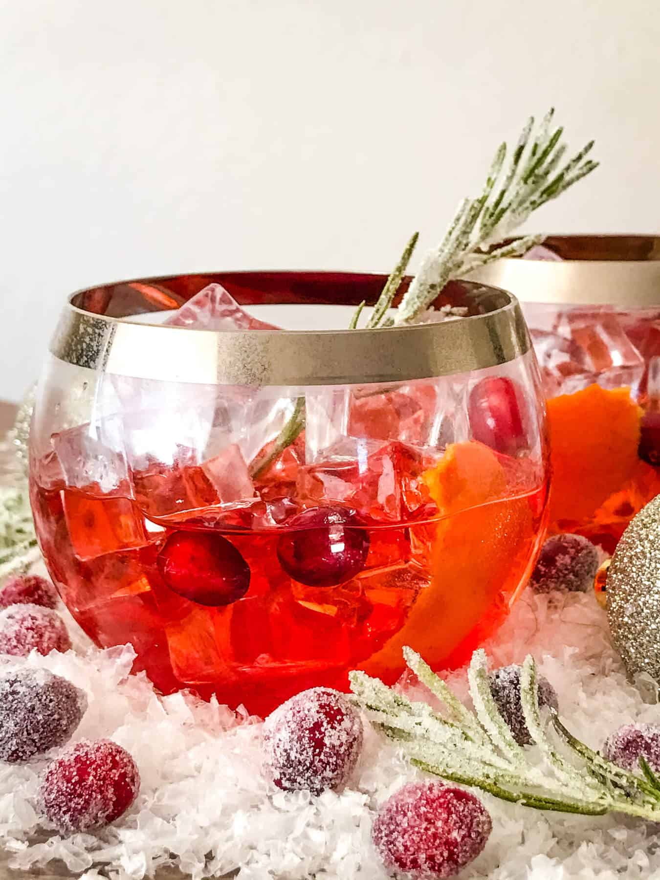 A glass of Cranberry Old Fashioned Cocktail with cranberries and rosemary in and around it
