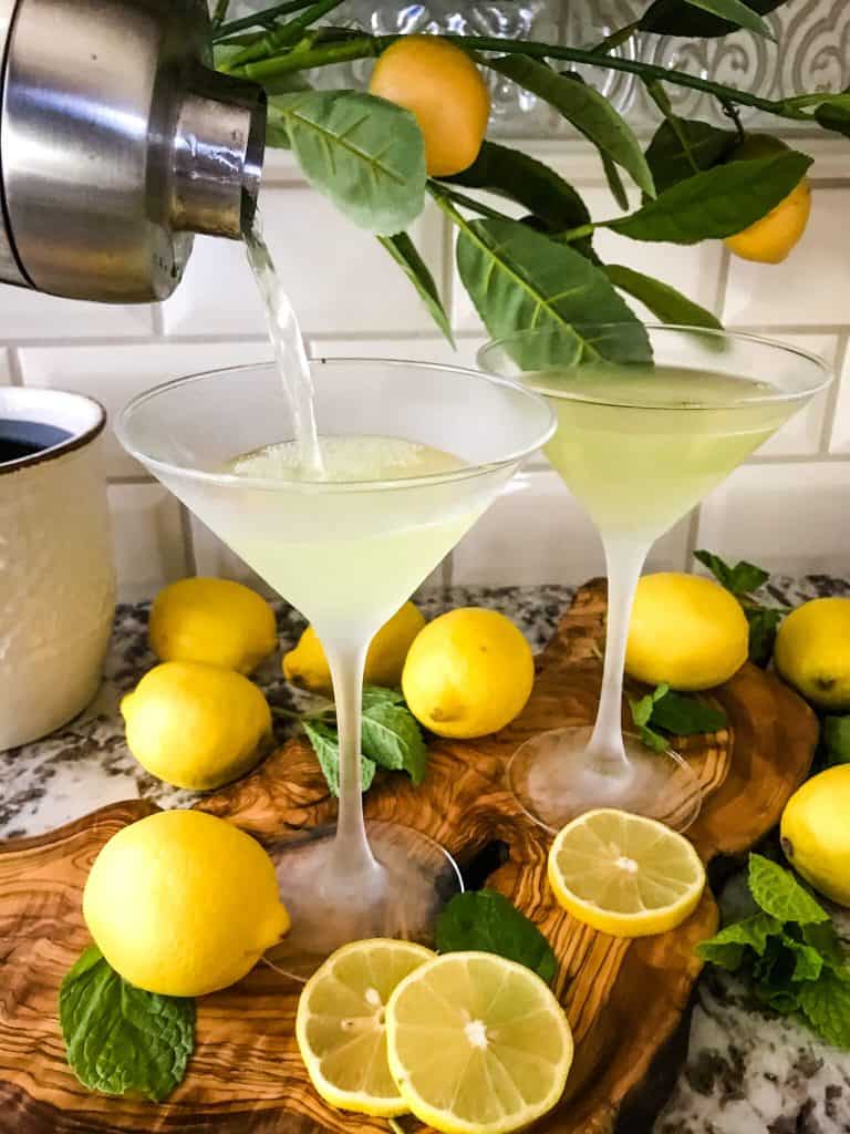 A cocktail shaker pouring a light yellow cocktail into glasses