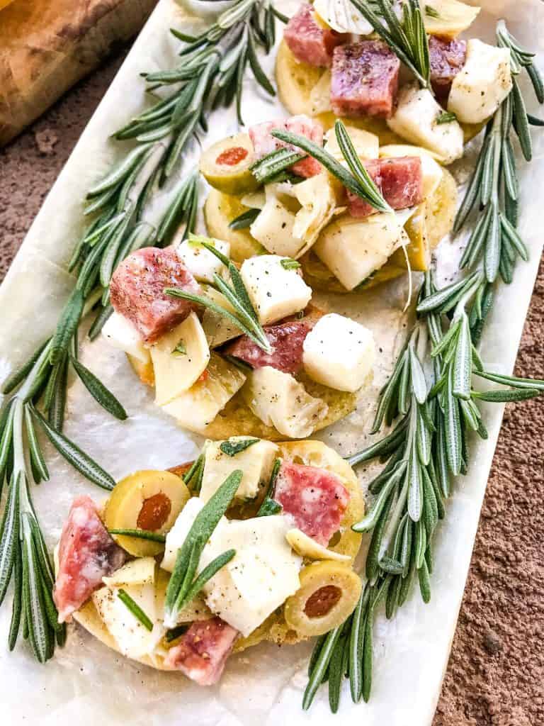 A row of Antipasti Crostini on a white platter with rosemary