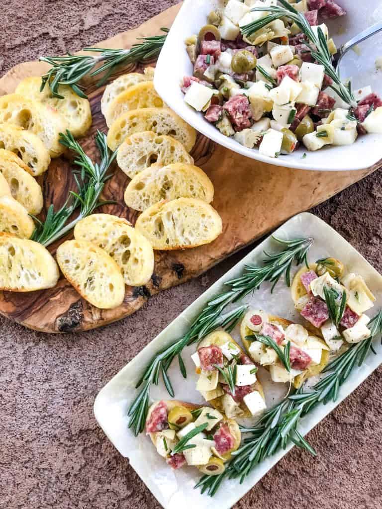 A wood platter holding the toasted bread slices and bowl of antipasti mixture with some topped bread slices next to them