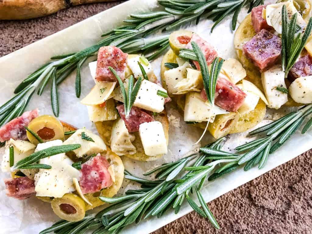 Slices of Antipasti Crostini lined up on a white platter with rosemary around them