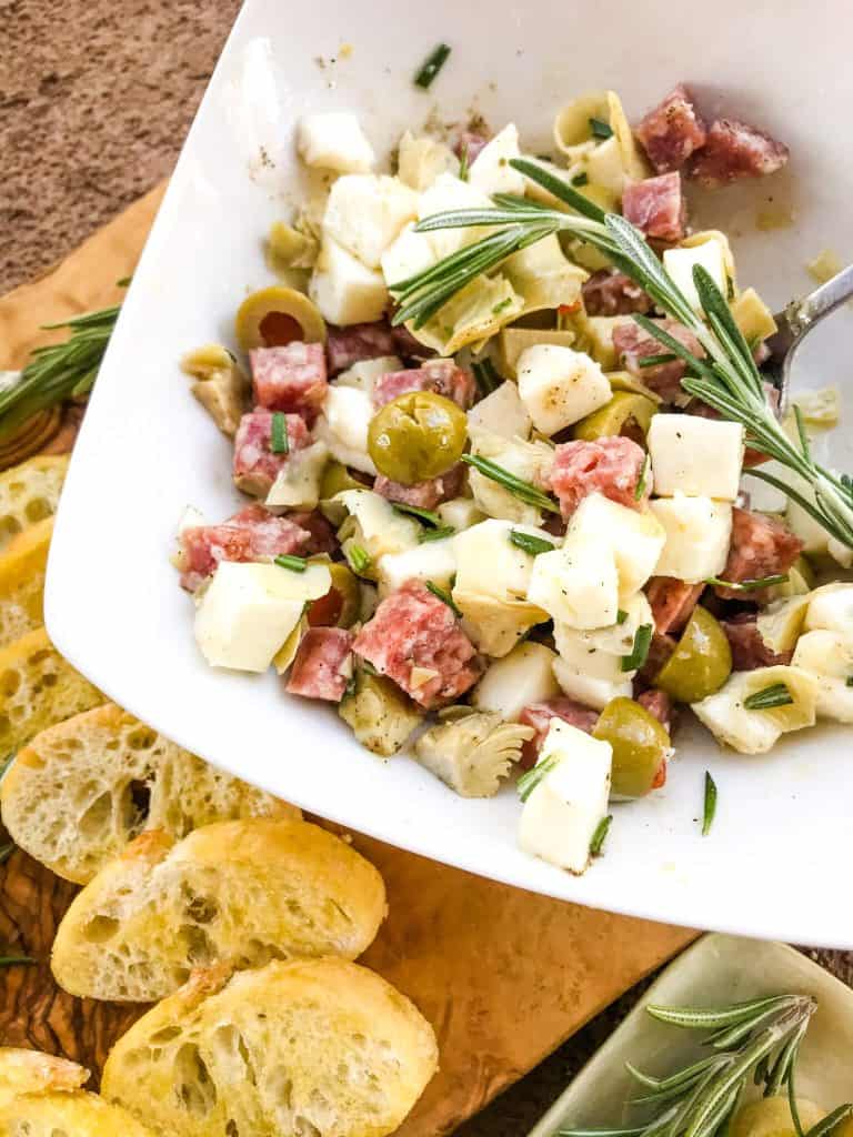 A bowl of salami, cheese, olives, and artichokes with bread slices next to it