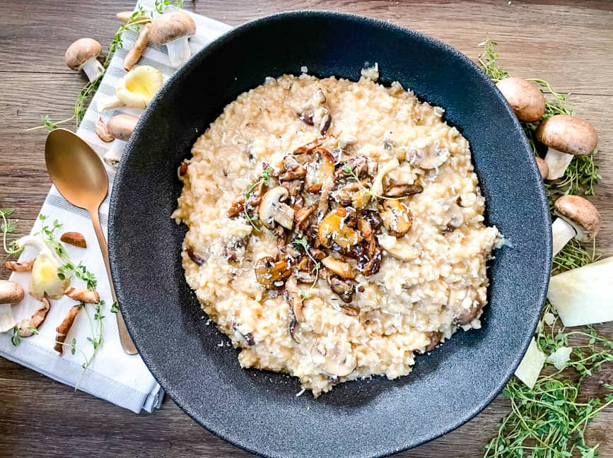 A big black bowl full of risotto from the top surrounded by mushrooms and thyme