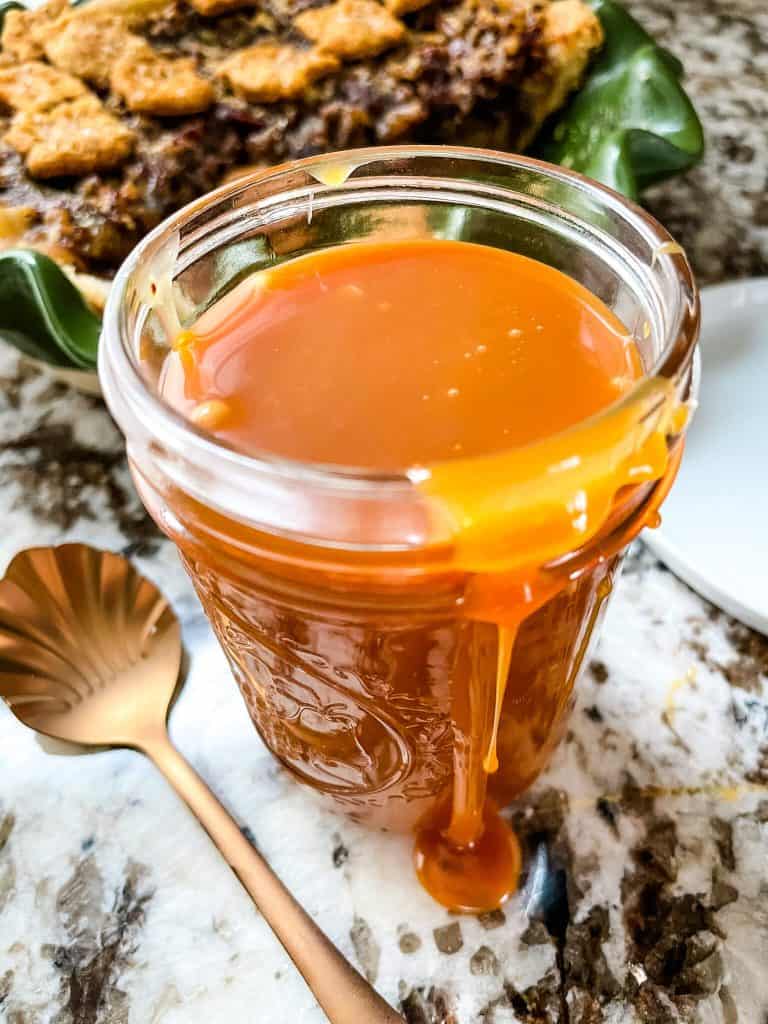 A glass jar of salted whiskey caramel sauce and a copper spoon in front of a pie