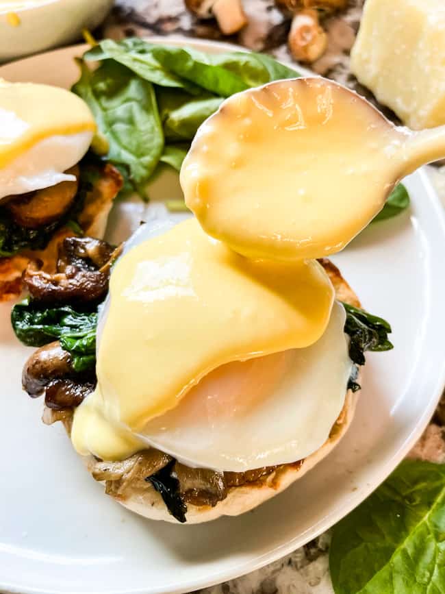 A spoon pouring hollandaise on a stack of Eggs Florentine Benedict