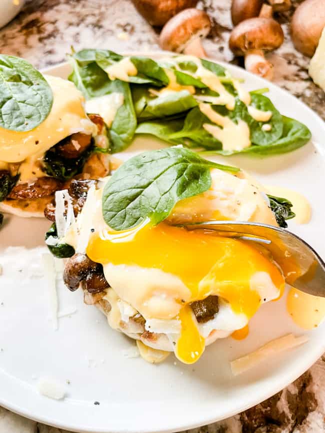 A fork breaking an egg stack open with the yolk running down the benedict