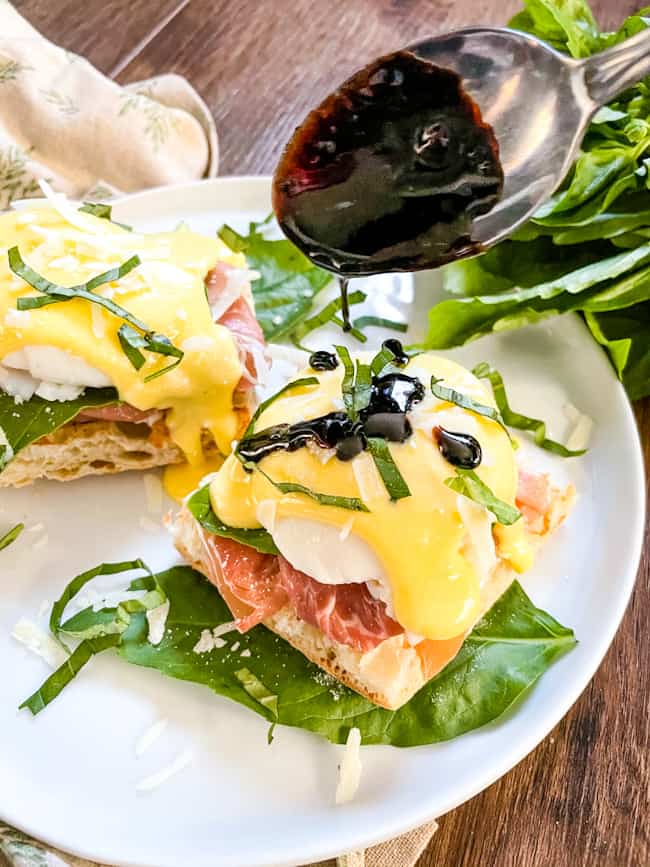 A spoon pouring balsamic glaze on a stack of Italian Prosciutto Eggs Benedict