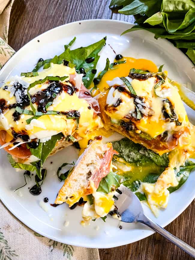 A fork with a piece of Italian Prosciutto Eggs Benedict on it next to the remaining stacks on a white plate