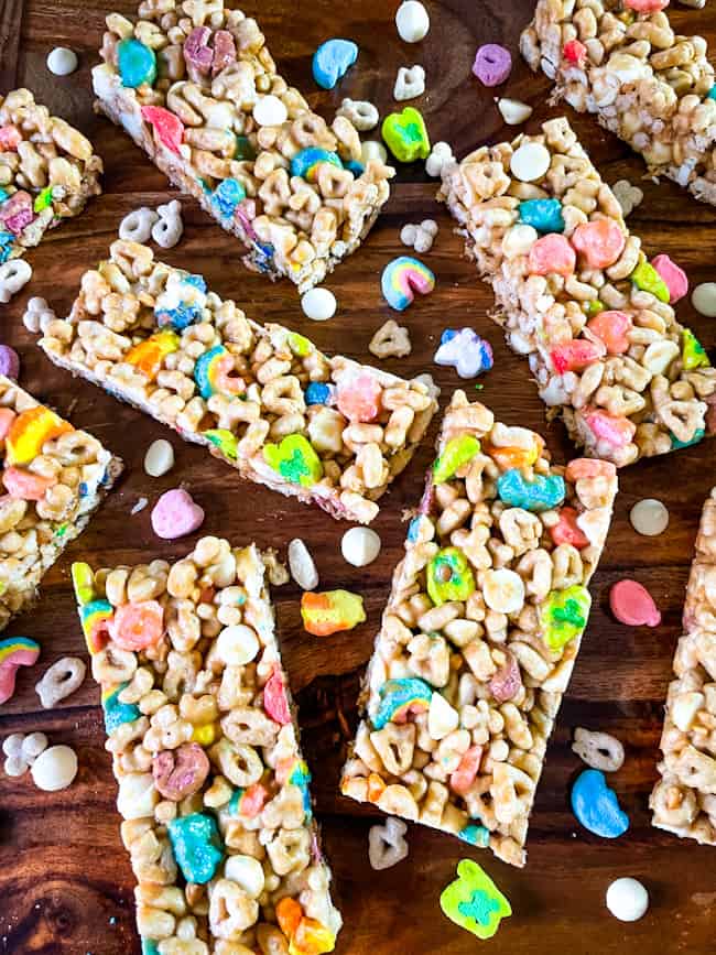 Lucky Charms Cereal Bars scattered on a wood surface with marshmallows and cereal around them