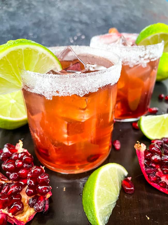 Two cocktails surrounded by limes and pomegranate pieces and seeds