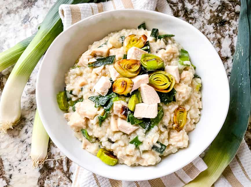 A white bowl of Chicken and Leek Risotto on a towel with leeks around it