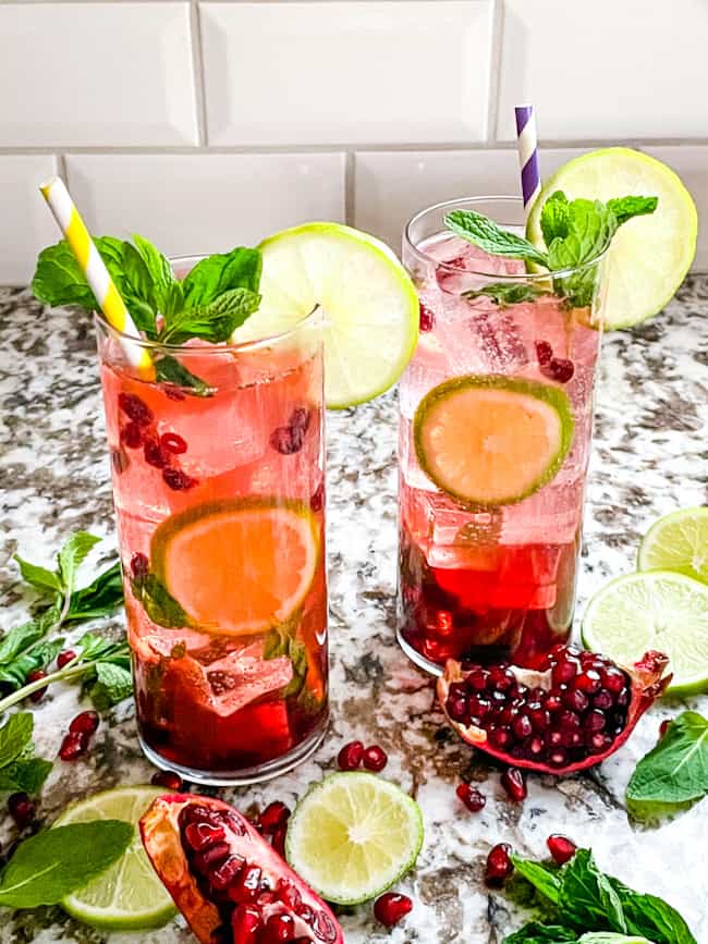 Two glasses of Pomegranate Mojito surrounded by pomegranates and limes