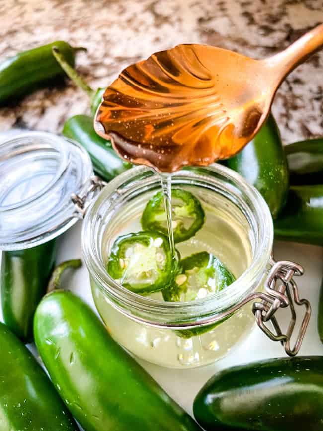 A copper spoon pouring Jalapeno Simple Syrup into a small glass jar with peppers around it