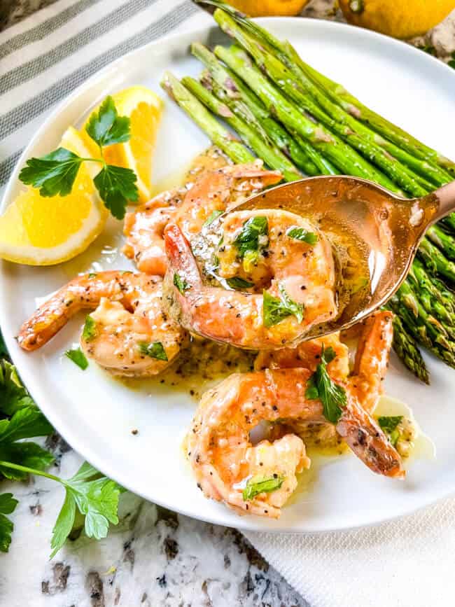 A copper spoon putting the seafood on a plate next to asparagus and lemon wedges