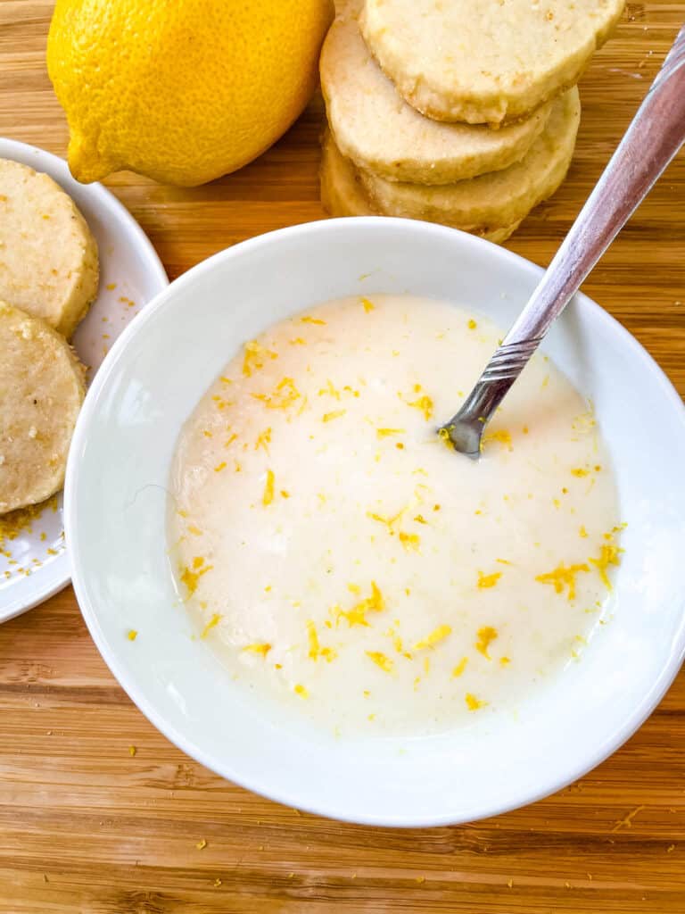 A spoon sitting in a white bowl full of Lemon Glaze on a cutting board