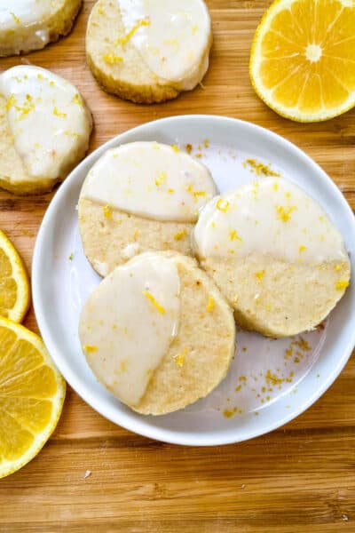 A white round plate with three Lemon Shortbread dipped in glaze surrounded by lemon slices and more cookies