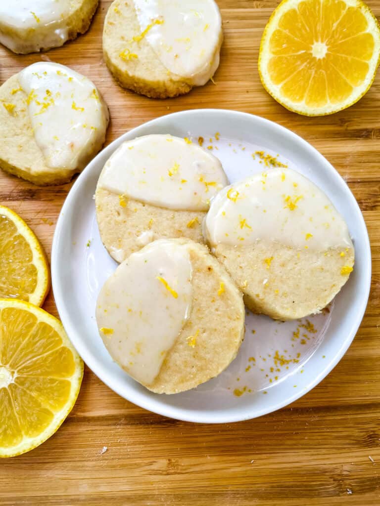 A white round plate with three Lemon Shortbread dipped in glaze surrounded by lemon slices and more cookies