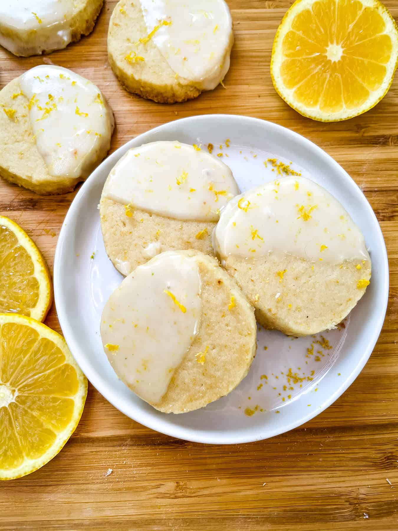 A white round plate with three Lemon Shortbread dipped in glaze surrounded by lemon slices and more cookies