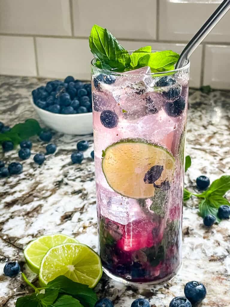 A tall glass of Blueberry Mojito with blueberries around it