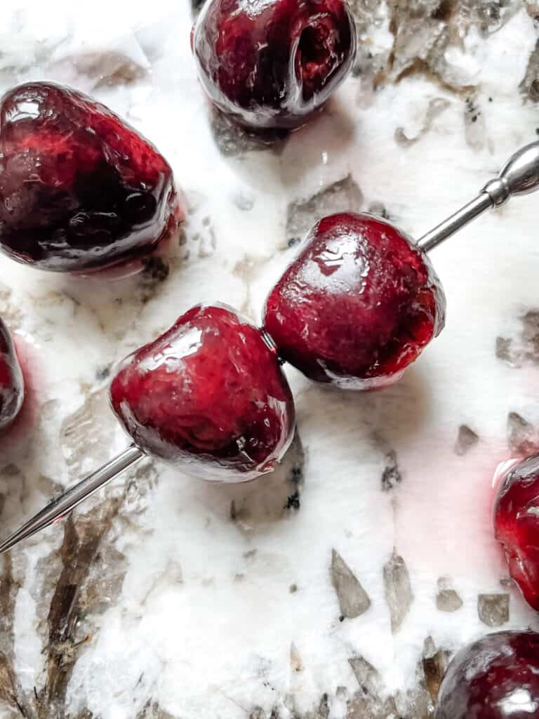 A cocktail skewer with two Brandied Cherries on a counter