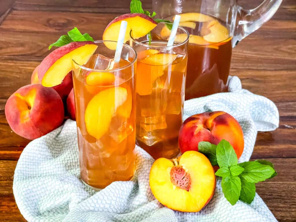 Two glasses of Peach Tea with a pitcher surrounded by peaches and mint