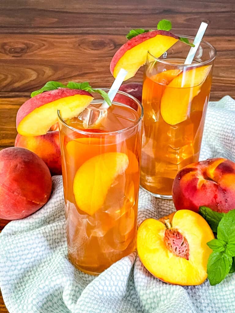 Two tall glasses full of Peach Tea with peaches in and around them