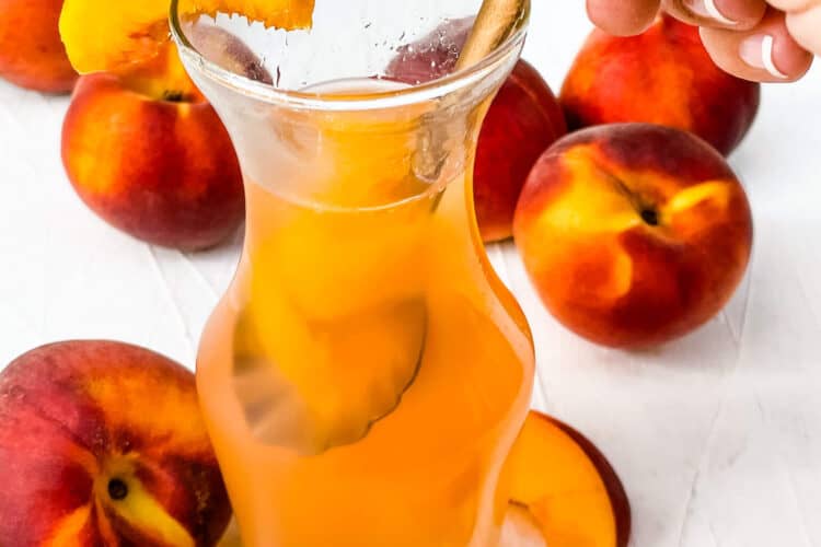 A hand dunking a spoon into a jar of Peach Syrup with peaches around it