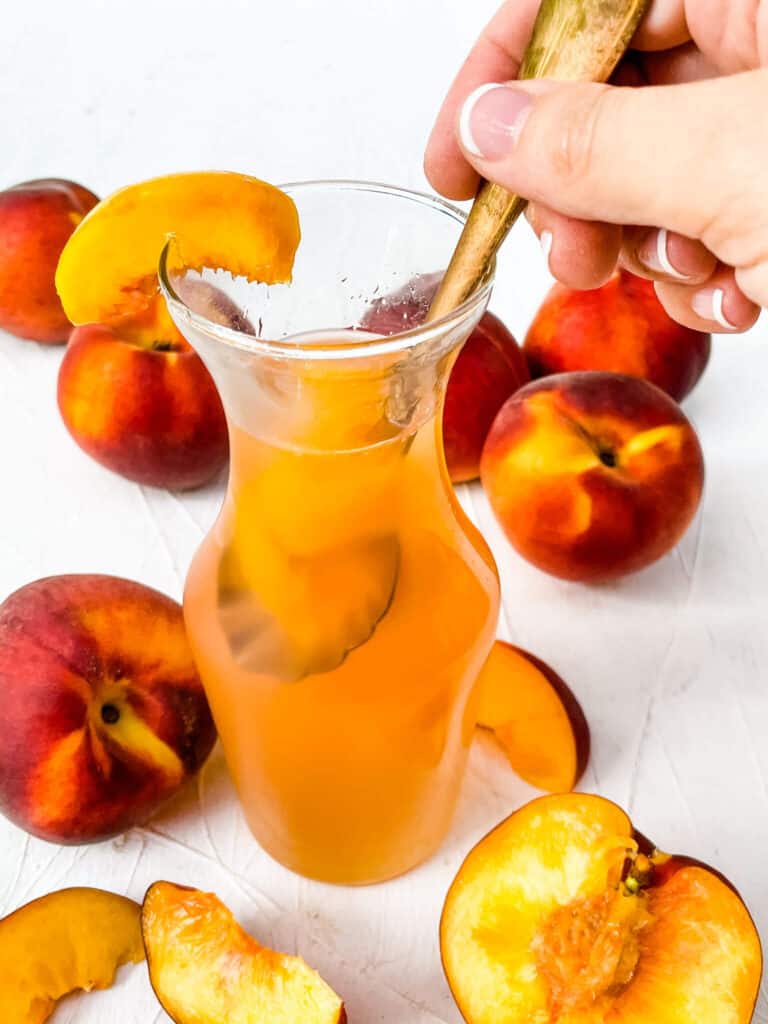 A hand dunking a spoon into a jar of Peach Syrup with peaches around it
