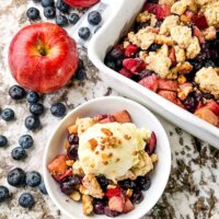 A serving of Apple and Blueberry Crumble in a bowl surrounded by blueberries and the serving dish nearby