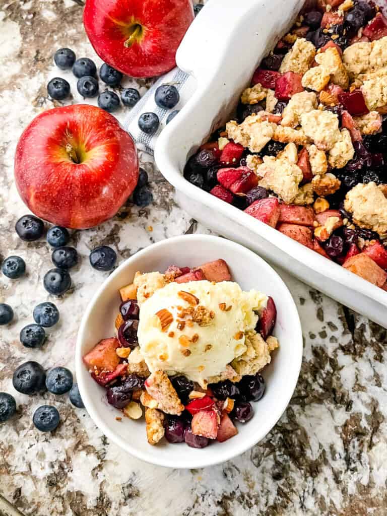 A serving of Apple and Blueberry Crumble in a bowl surrounded by blueberries and the serving dish nearby