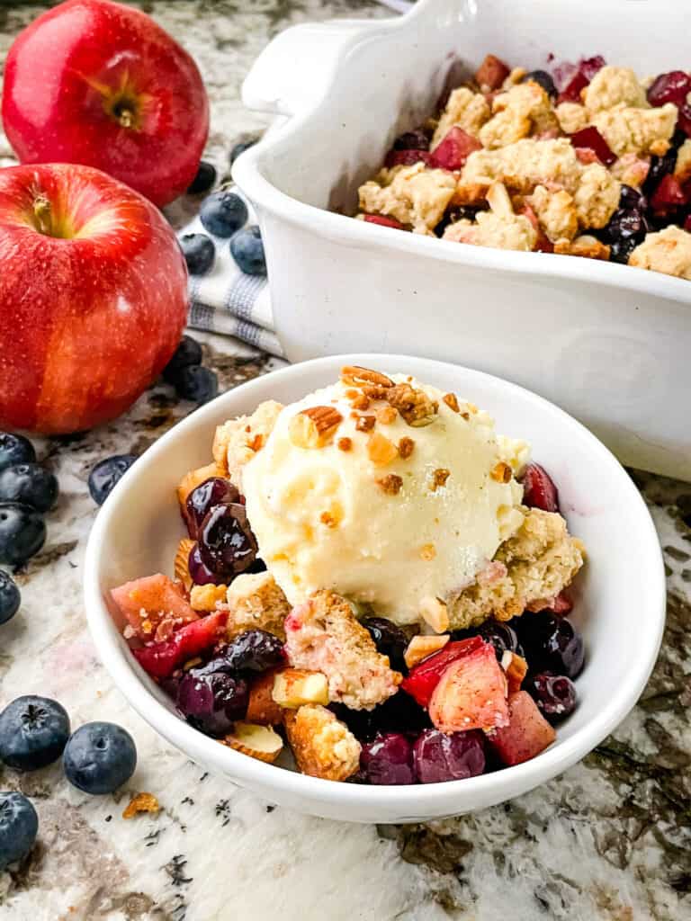 A serving of the Apple and Blueberry Crumble in a white bowl with the serving dish in the background