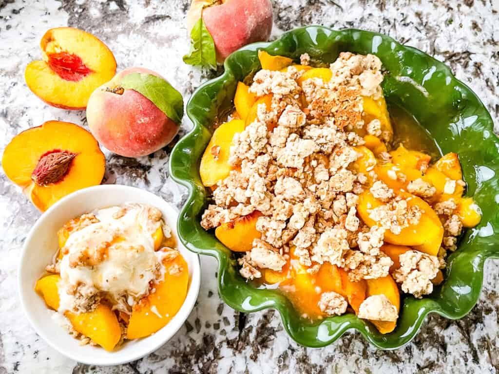 Top view of the Peach Crumble in a green pie dish with a small portion in a white bowl next to it. Peaches around the dishes.