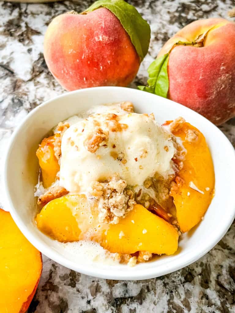 A small white bowl filled with a portion of Peach Crumble topped with vanilla ice cream