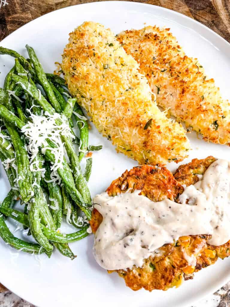 A white plate with Parmesan Air Fried Chicken Tenders, Corn and Zucchini Fritters, and Air Fried Green Beans with white gravy.